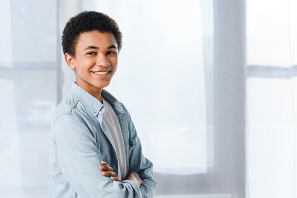 uśmiechający się Afryki amerykański nastolatek stojąc ze skrzyżowanymi ramionami i patrząc na kamery w domu - Zdjęcie, obraz