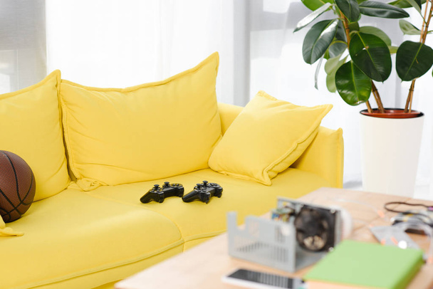 gamepads y pelota de baloncesto en sofá amarillo en casa
 - Foto, Imagen