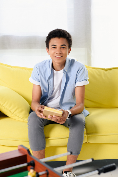 mosolyogva ült otthon sárga kanapén könyv afro-amerikai tinédzser - Fotó, kép