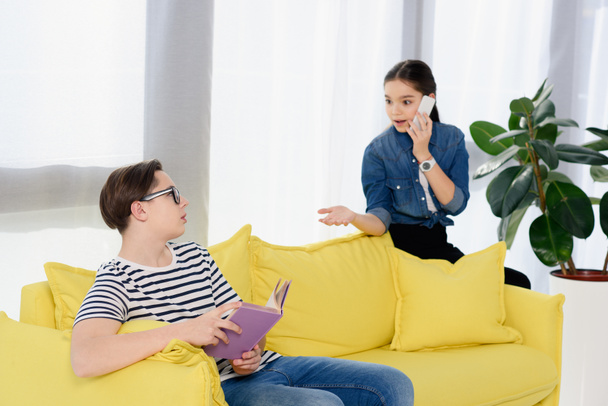 teenager talking by smartphone and looking at boy sitting on sofa with book at home - Foto, Bild