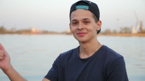 Young man smiles and makes a finger gun gesture on the Dnipro bank in slo-mo                            Portrait of a cheerful young man in a baseball cap showing a finger gun gesture and smiling like a mischief on the Dnipro bank in spring in slo-mo - Filmagem, Vídeo