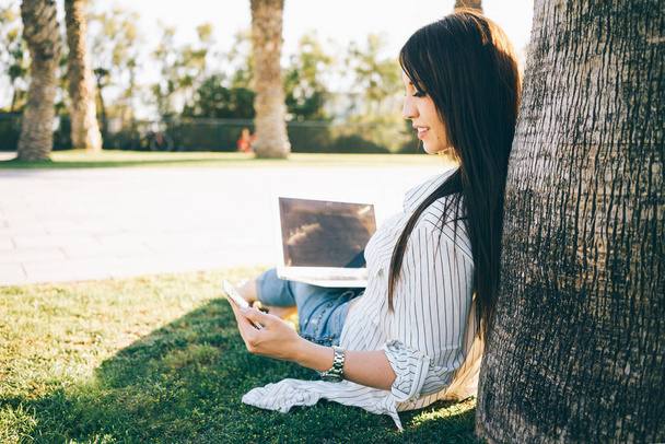 Young woman in a striped shirt with laptop on her knees is using smartphone while relaxing in the city garden. - Fotoğraf, Görsel