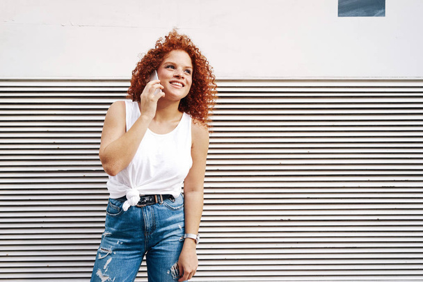 A beautiful young woman with red hairs talking on a smartphone while standing beside the wall on a street  - Photo, Image