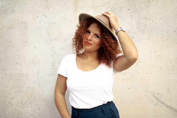 charming red haired female woman with a freckled face wearing a white t-shirt and a hat on the concrete background - Photo, Image