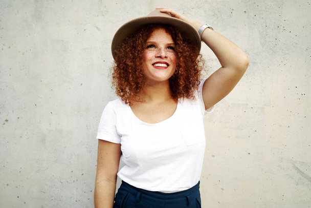 charming red haired female woman with a freckled face wearing a white t-shirt and a hat on the concrete background - Photo, Image