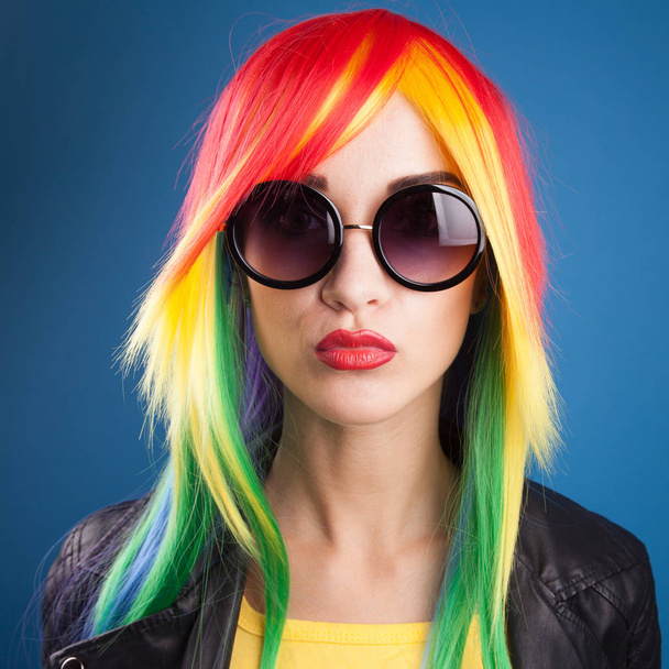 beautiful young woman wearing color wig and sunglasses against blue background - Фото, изображение