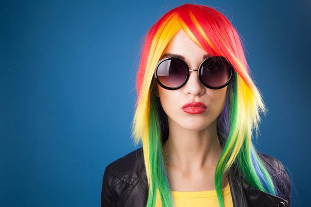 beautiful young woman wearing color wig and sunglasses against blue background - Photo, Image