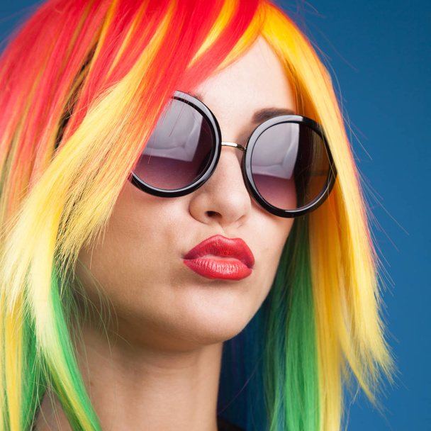 beautiful young woman wearing color wig and sunglasses against blue background - Foto, Bild