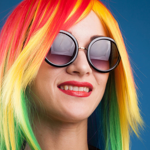 beautiful young woman wearing color wig and sunglasses against blue background - Фото, изображение