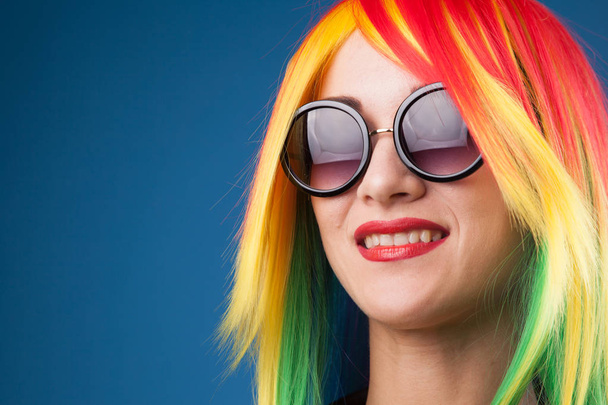 beautiful young woman wearing color wig and sunglasses against blue background - Foto, afbeelding