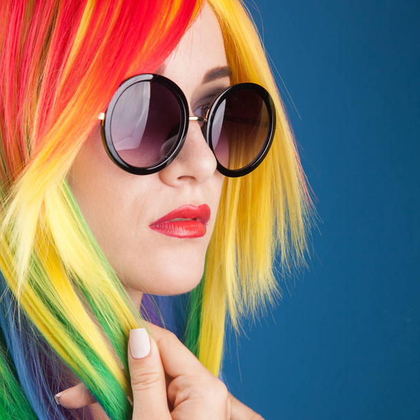 beautiful young woman wearing color wig and sunglasses against blue background - Zdjęcie, obraz