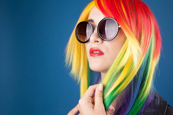 beautiful young woman wearing color wig and sunglasses against blue background - Fotoğraf, Görsel