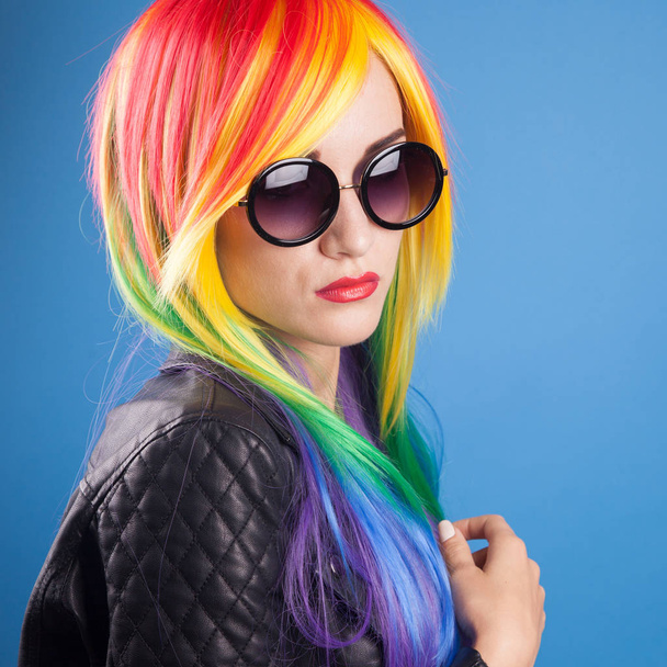 beautiful young woman wearing color wig and sunglasses against blue background - Fotó, kép