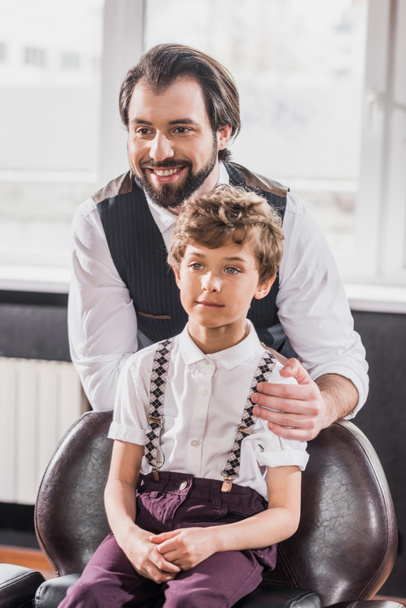 очаровательный маленький ребенок сидит на стуле в парикмахерской пока счастливый парикмахер стоит позади него
 - Фото, изображение
