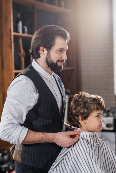 красивый парикмахер смотрит на отражение маленького ребенка, сидящего в кресле в парикмахерской
 - Фото, изображение