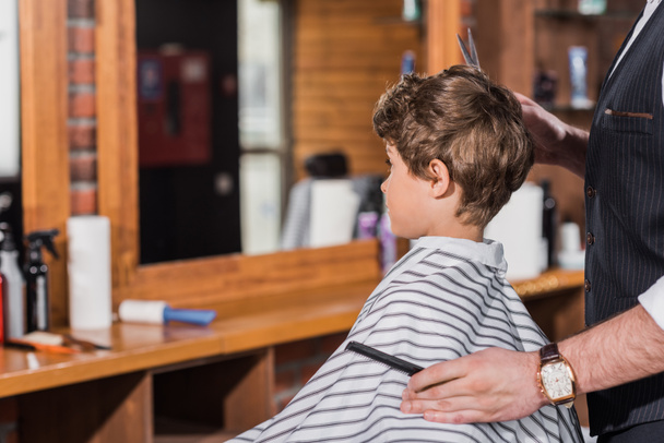 little curly kid covered with striped cloth sitting at barbershop while barber cutting his hair - Foto, Imagem