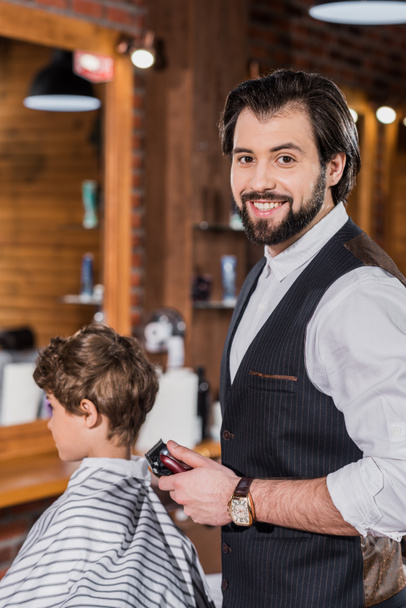 barber cutting hair of little kid with Hair Clipper and looking at camera - 写真・画像