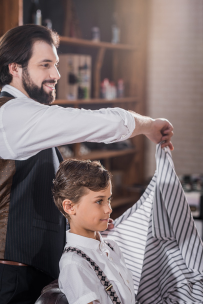 smiling barber covering adorable little kid with striped cloth at kids barbershop - Photo, Image