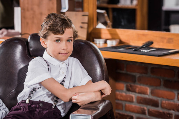 smiling little kid sitting in barber chair at barbershop and looking at camera - Foto, immagini