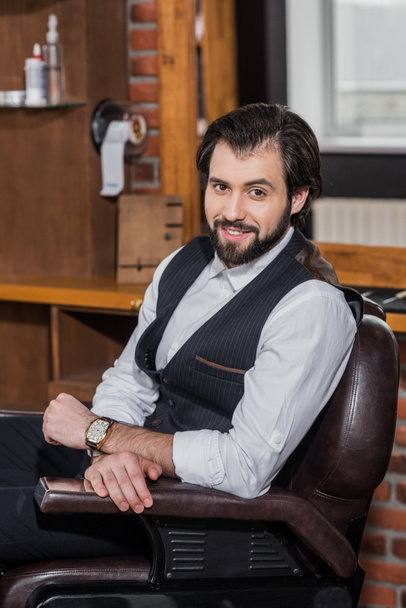 happy barber sitting on chair at workplace and looking at camera - Foto, Bild