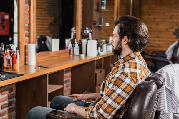 knappe bebaarde man in plaid shirt zit op stoel bij barbershop - Foto, afbeelding