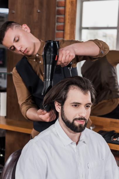 σοβαρή κουρέας χτενίζει τα μαλλιά του πελάτη με στεγνωτήρα μαλλιών στο κουρείο - Φωτογραφία, εικόνα