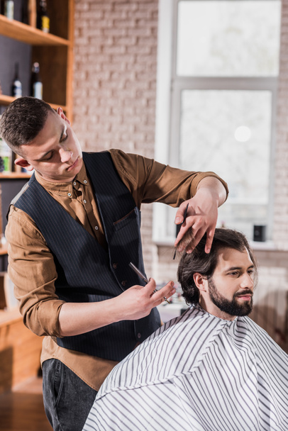 bearded young man covered with striped cloth getting haircut from professional barber at barbershop - Foto, Bild