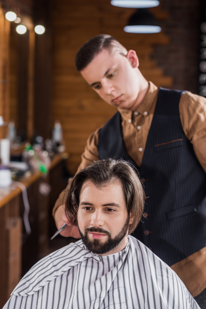 bearded young man getting haircut from professional barber at barbershop - Photo, Image