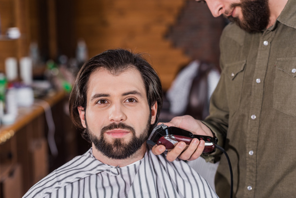 Friseur rasiert glücklich gutaussehenden Kunden mit Haarschneidemaschine - Foto, Bild