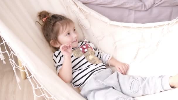 Little smiling girl chilling and swinging on a white hammock. Indoors - Imágenes, Vídeo