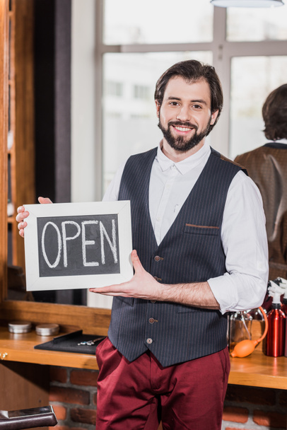 happy bearded barber holding open signboard in front of workplace - Photo, Image