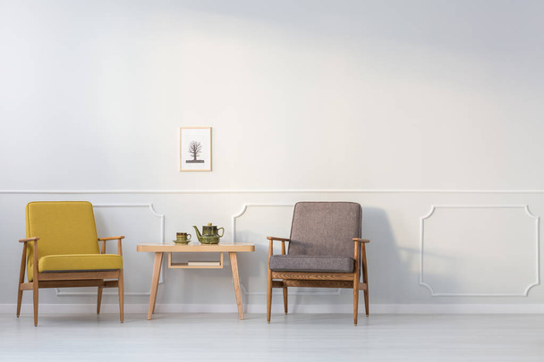 Wooden table between yellow and grey armchair against white wall with poster in living room interior - Foto, Imagem