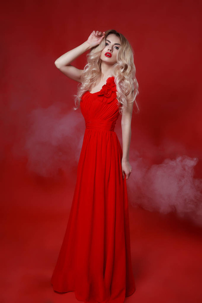 young beautiful girl with long blond hair. woman with curly hair. smoke on the red background. lady in long red dress isolated on red background - Photo, Image
