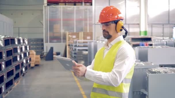 Een mannelijke werknemer werkt met zijn gadget in een magazijn, het dragen van speciale hoed en bril. 4k. - Video