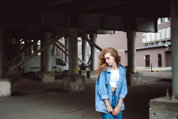 street portrait of a young attractive emotional girl with curly slips dressed in a trendy blue jeans suit on a style walking in the open air - 写真・画像