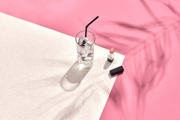 Hochwinkelblick auf Lippenstift und Trinkglas mit Eis auf dem Tisch. rosa und weißer Hintergrund mit Schatten von einem Palmblatt - Foto, Bild