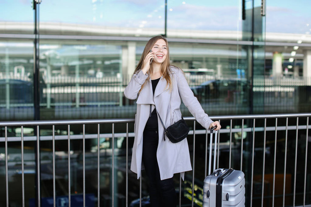 Красивая женщина, стоящая с валиком и разговаривающая по смартфону возле аэропорта
. - Фото, изображение