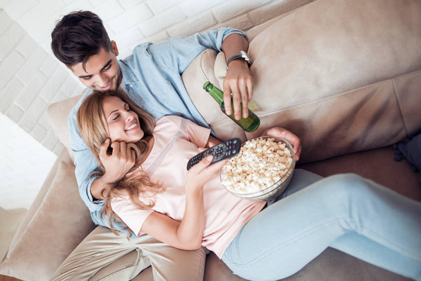 Young couple relaxing in their apartment, lying on the couch, watching a movie and eating popcorn. - Photo, image