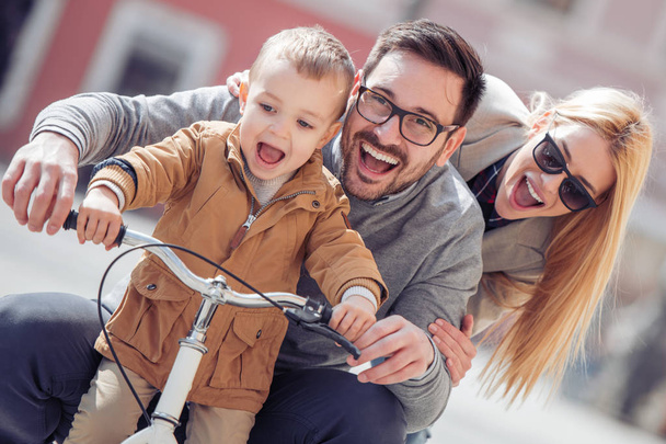 Father and mother teaching their son to ride bicycle in city on sunny day. - Zdjęcie, obraz