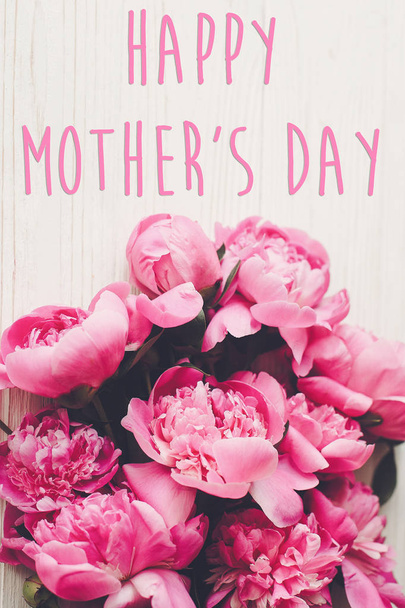 glücklicher Muttertagstext auf rosa Pfingstrosen Strauß auf rustikalem weißem Holzhintergrund, Draufsicht. Florales Grußkartenkonzept, flach gelegt. Muttertag. vertikales Federbild - Foto, Bild