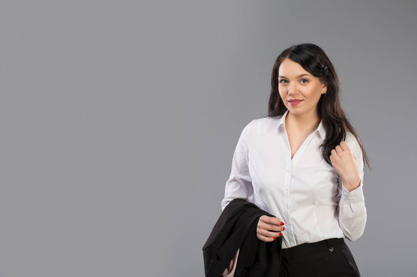 Portret van een mooie jonge zakenvrouw permanent met hand houden jas achter haar schouder tegen de grijze achtergrond en poseren modieus. Kijken naar camera. - Foto, afbeelding