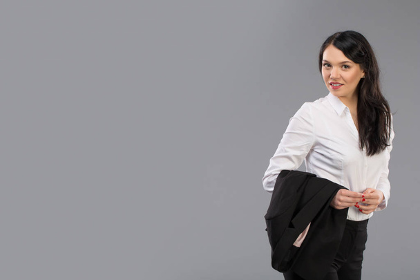 Portrait of a beautiful young business woman standing with hand holding jacket behind her shoulder against grey background and posing fashionable. Looking at camera. - Photo, image