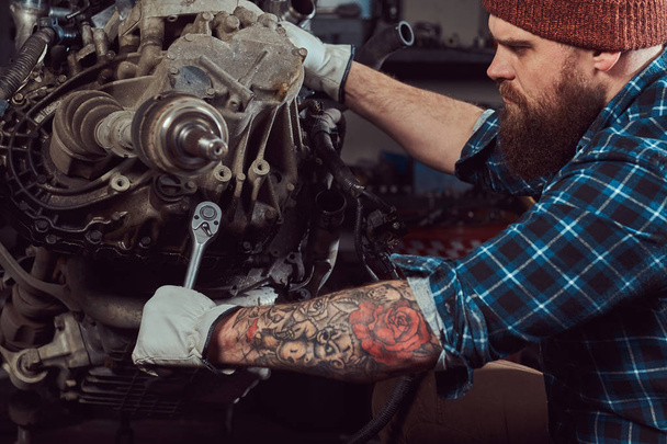 残忍な入れ墨ひげを生やしたメカニックの専門家は、ガレージに油圧リフトに発生する車のエンジンを修復します。サービス ステーション. - 写真・画像