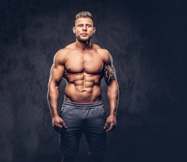 A handsome shirtless tattooed bodybuilder with stylish haircut and beard, wearing sports shorts, posing in a studio. Isolated on a dark background - Фото, изображение