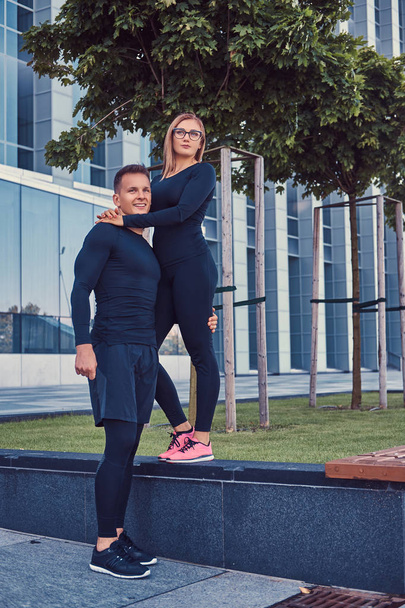 Happy fitness couple in a sportswear, sexy blonde girl and a handsome muscular guy stands in the modern city against a skyscraper. - 写真・画像