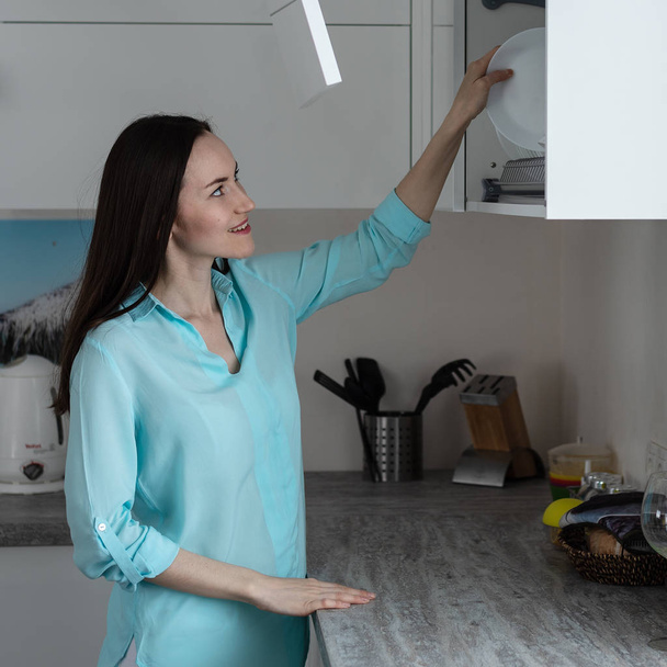 Jovem dona de casa coloca pratos limpos na prateleira do rack de secagem no interior da cozinha branca, conceito de tarefas domésticas
 - Foto, Imagem