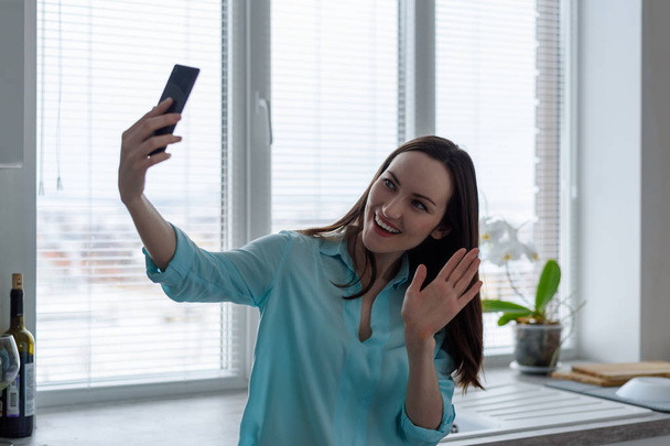 Een jonge brunette communiceert online via een smartphone in de keuken, een groet symbool zwaaien - Foto, afbeelding