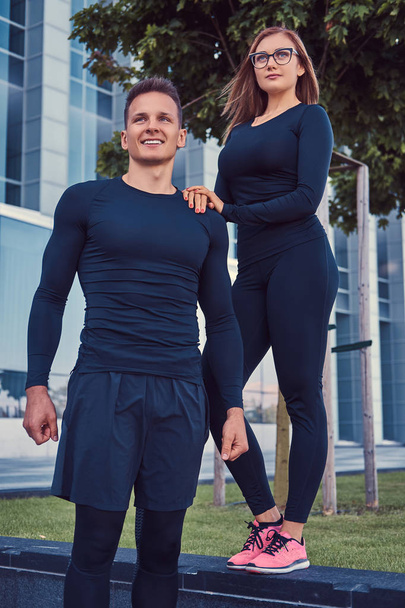 glückliches Fitness-Paar in Sportbekleidung, sexy blondes Mädchen und ein gutaussehender muskulöser Kerl steht in der modernen Stadt vor einem Wolkenkratzer. - Foto, Bild