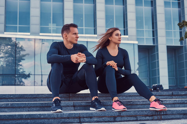 Una joven pareja de fitness en ropa deportiva, se sienta en los escalones de la ciudad moderna contra un rascacielos. concepto de estilo de vida saludable
. - Foto, Imagen