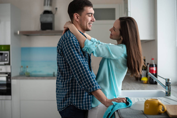 Jovem casal feliz abraçando e olhando uns para os outros no interior de uma nova cozinha, felicidade em uma nova casa
. - Foto, Imagem